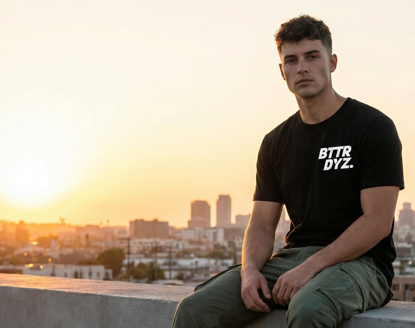 Man sitting on a rooftop with a cityscape in the background during sunset, wearing a black t-shirt with 'BTTR DYZ' text.