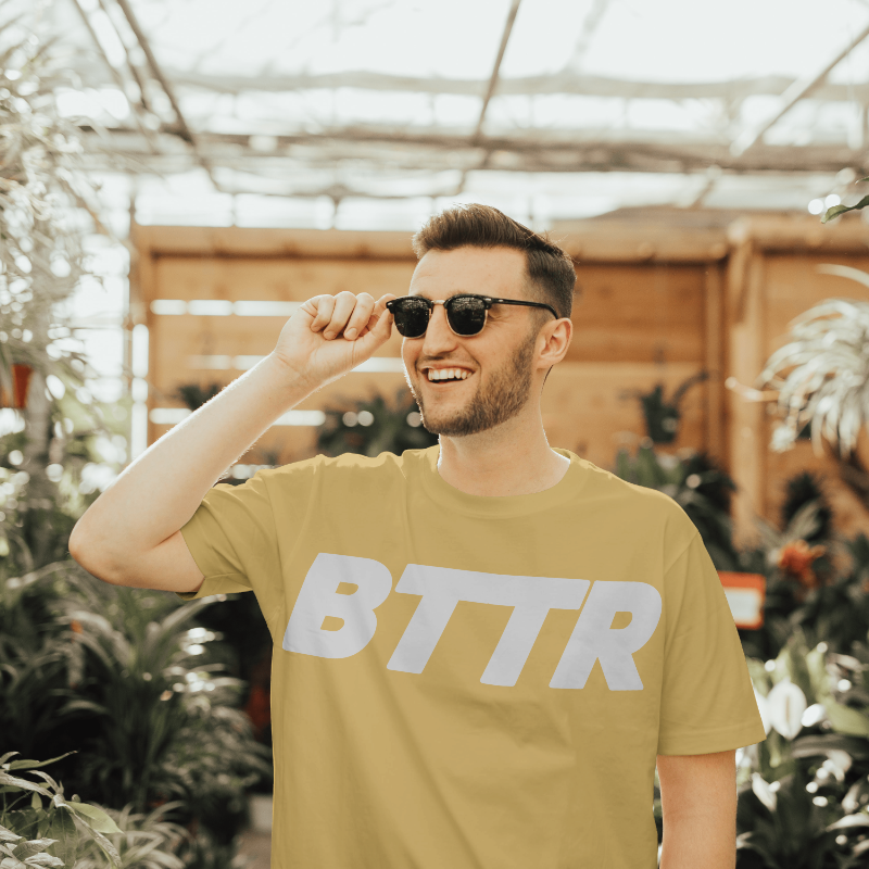 Man wearing a mustard yellow t-shirt with 'BTTR' on it, standing in a greenhouse.